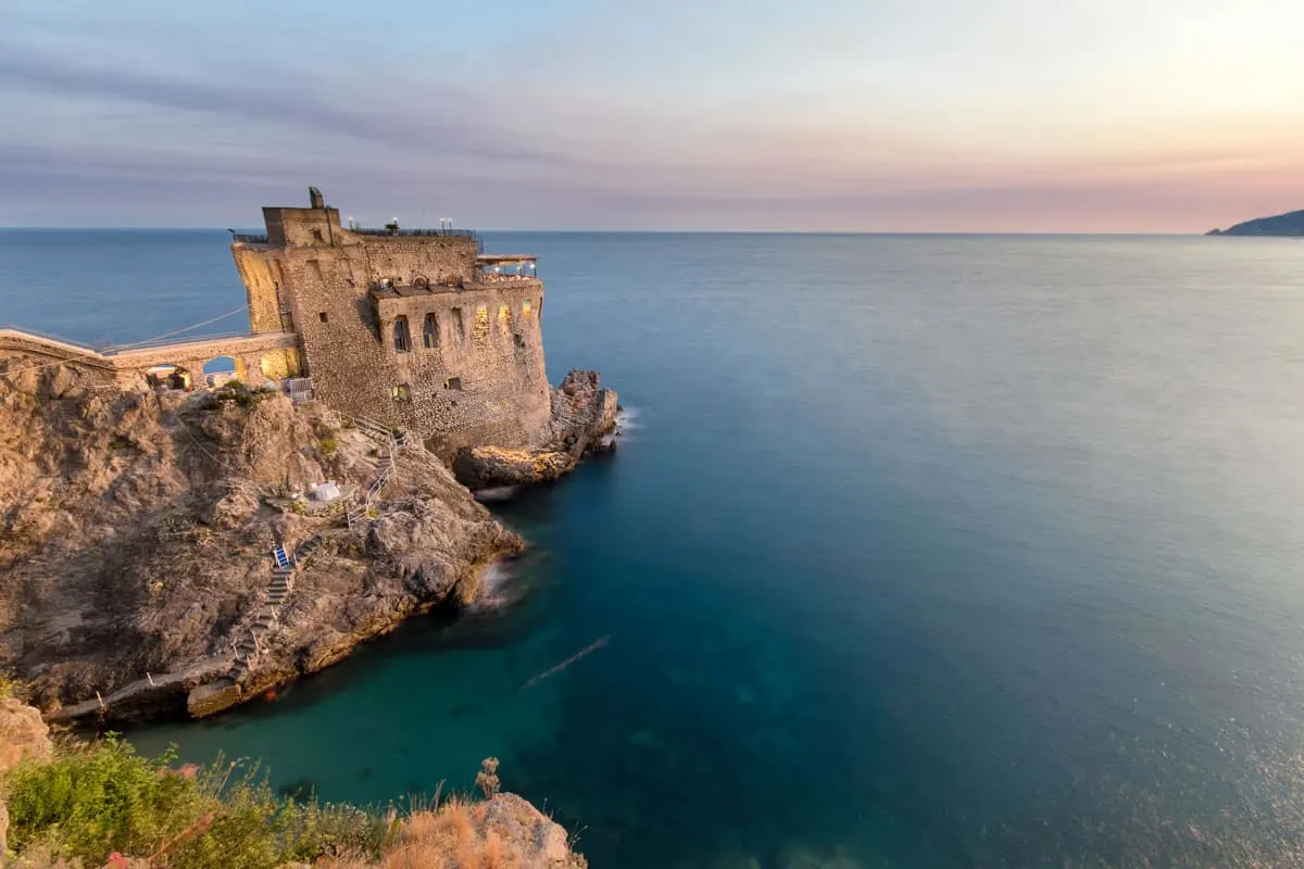 A fort juts out over the ocean at dusk. The lights from the restaurant at the top of the fort glow.