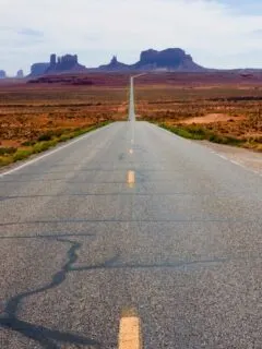 A lone car driving down a desert road at dusk with rock formations in the distance.