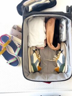 An open suitcase on a white bed with a few items packed and some on the bed. It demonstrates how to pack shoes for travel.