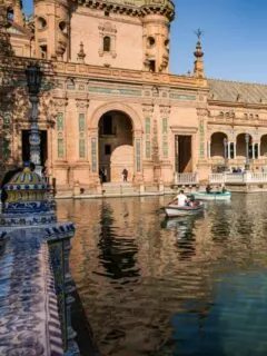 People in row boats on a small river running past ornate historical Spanish buildings in the south of Spain.