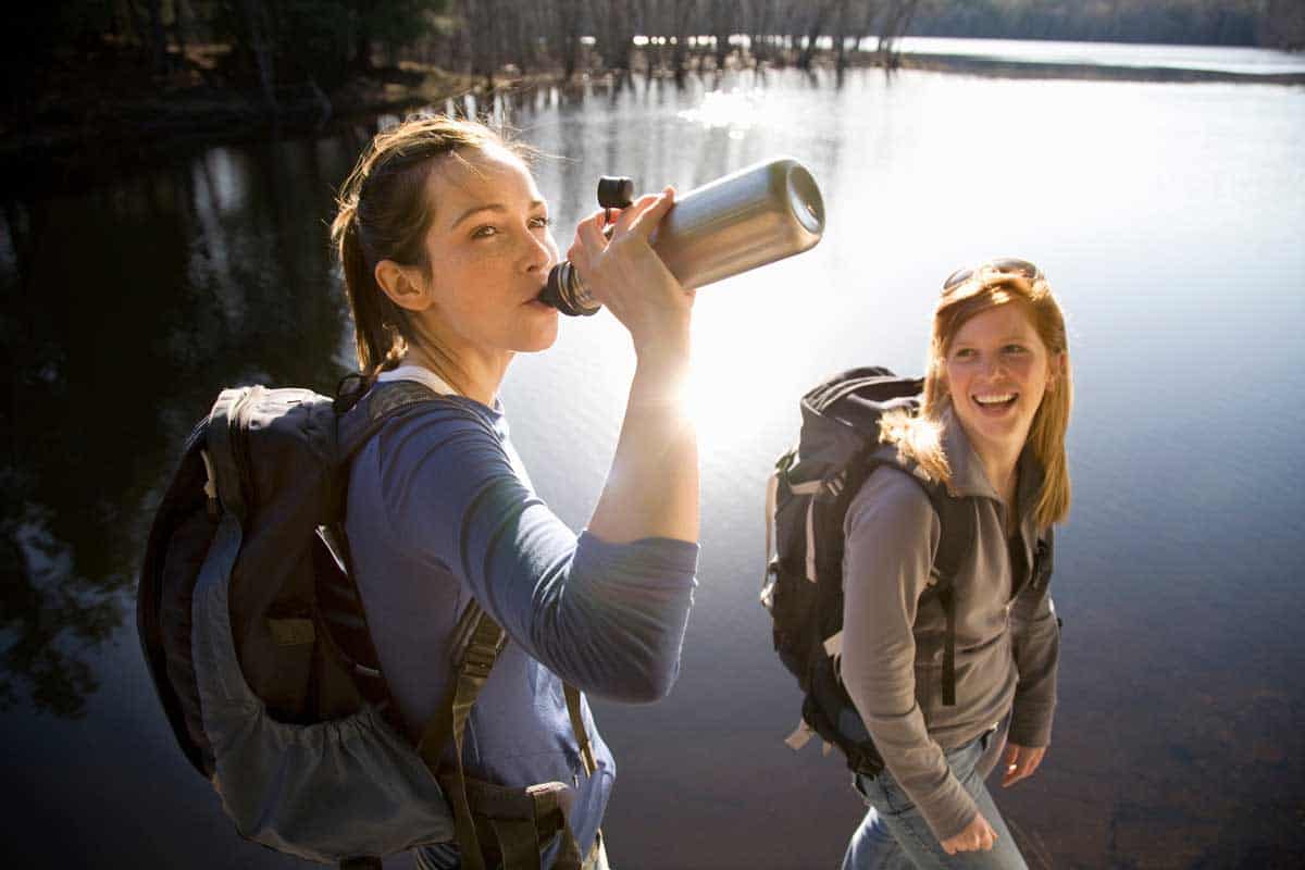 Two female hikers sharing water next to a lake. 