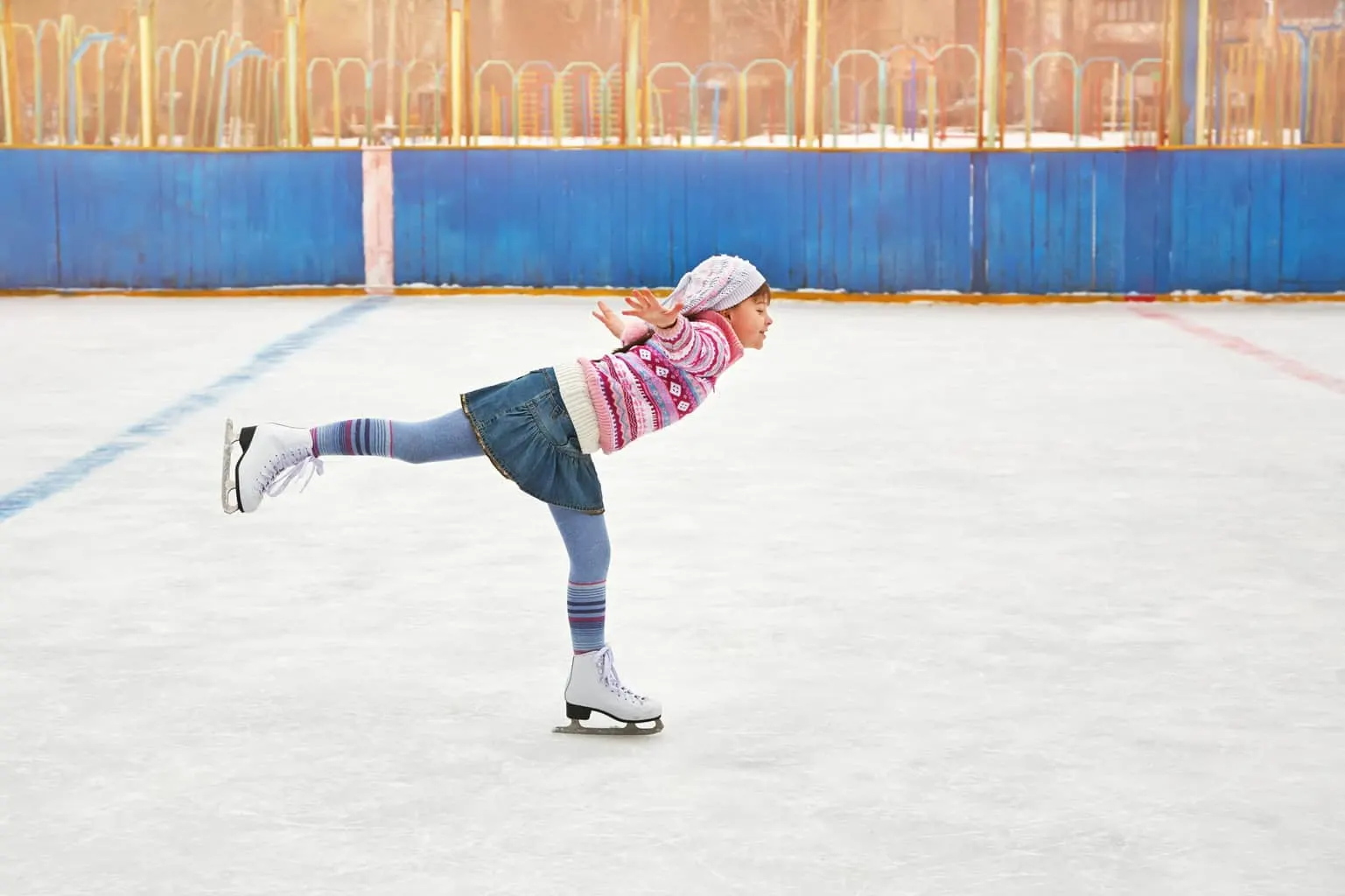 A girl in a patterned jacket and knit hat gliding on one skate at an outdoor rink