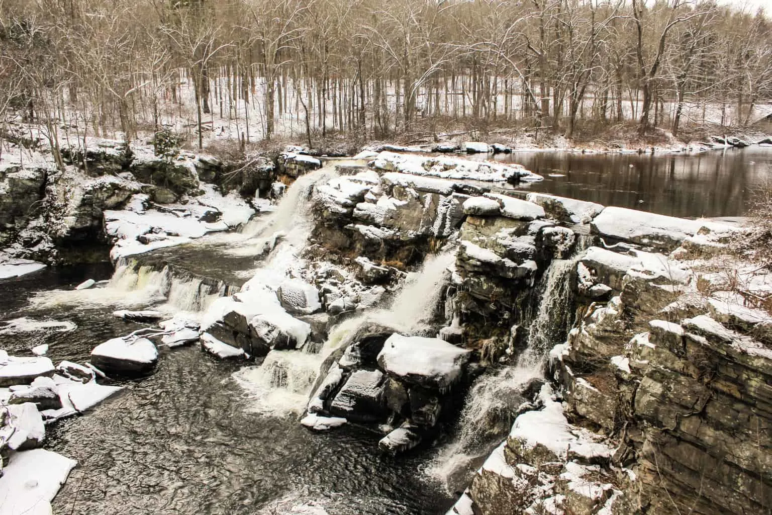 A partly frozen river spilling over rock ledges with snow-covered banks and bare trees in the Poconos in winter