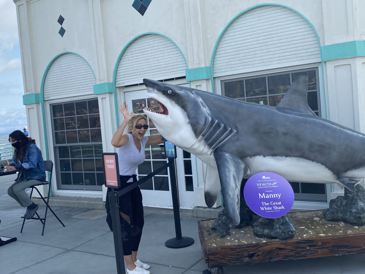 woman standing under pretend shark at Hunington Beach in California. Her arms are up an waving under the shark