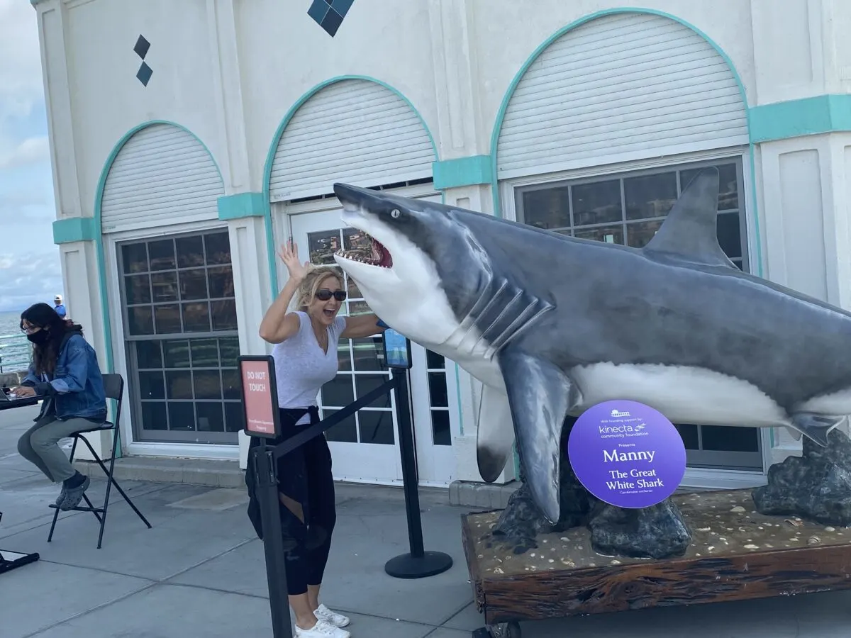 woman standing under pretend shark at Hunington Beach in California. Her arms are up an waving under the shark