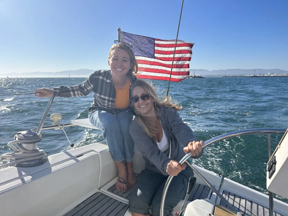 Two women smiling on a sailboat in the Pacific Ocean with the American Flag in the background