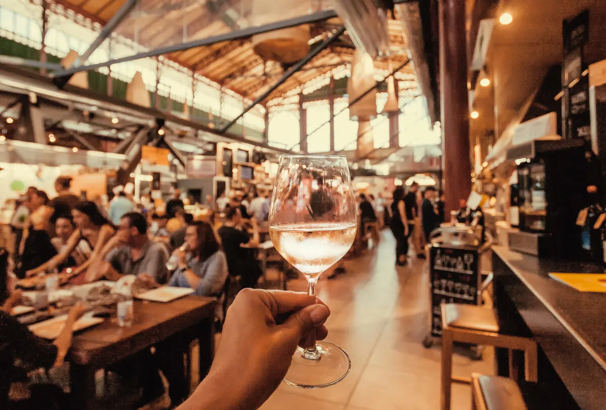 Hand holding up a glass of white wine in a busy restaurant with many diners in the background