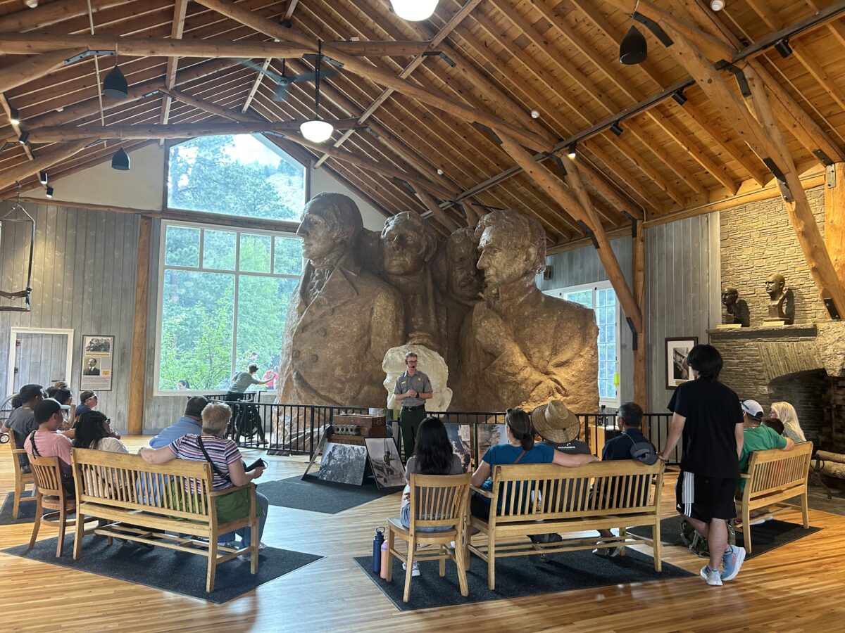 Guide giving a talk about the history of Mount Rushmore in a small building at the base of Mount Rushmore. People sitting in benches and chairs listening