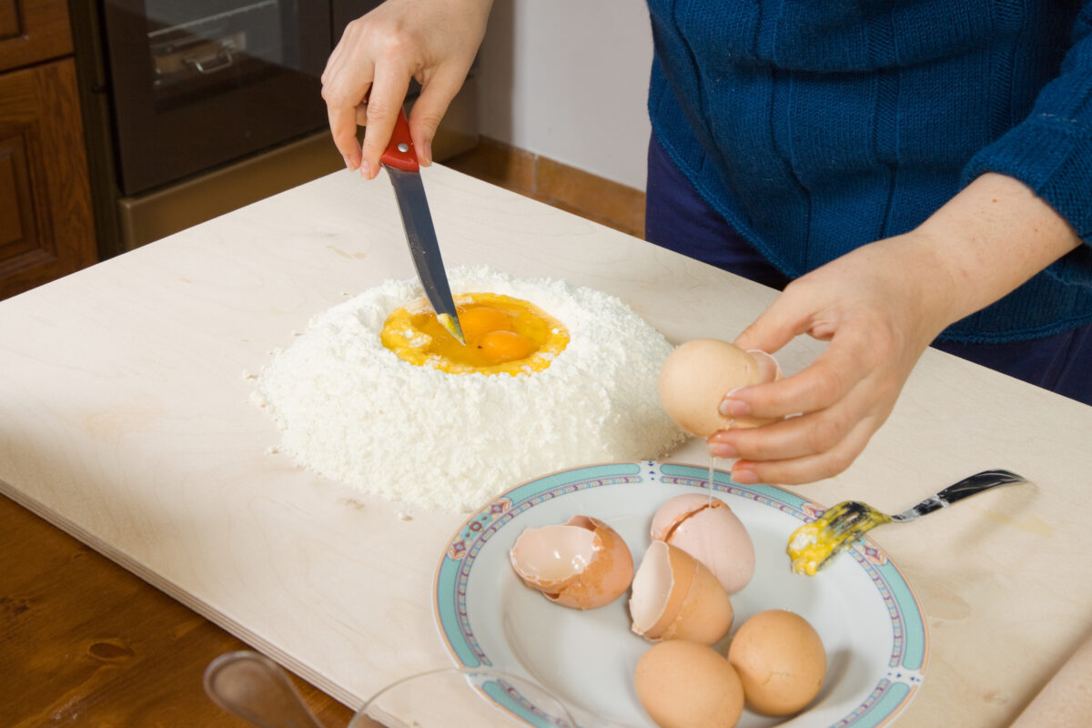 woman making pasta. Raw eggs sitting in a hole of flour waiting to be broken and mixed with the knife she is holding. Broken egg shells in a bowl on the side.