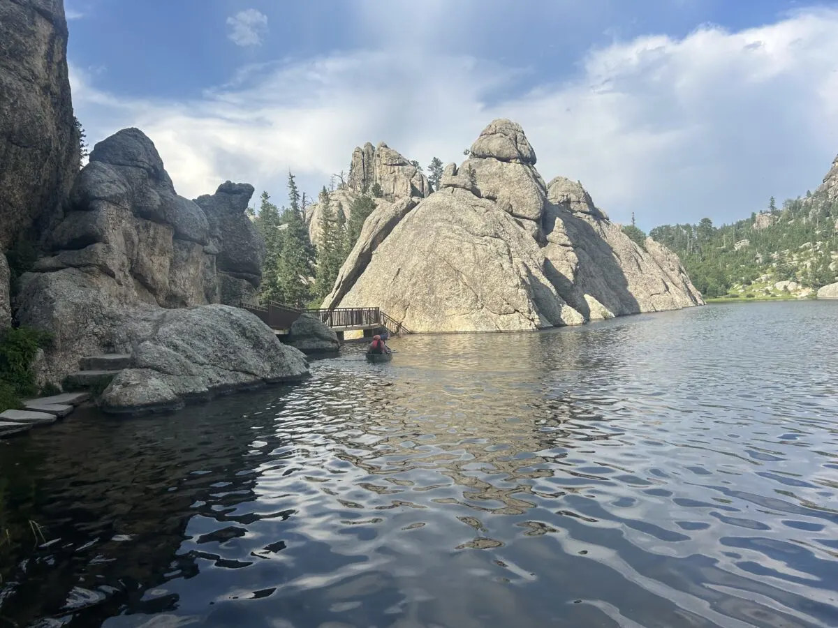 View of Sylvan Lake with large rocks  in the background and person kayaking on a sunny day in Custer Stae Park South Dakota