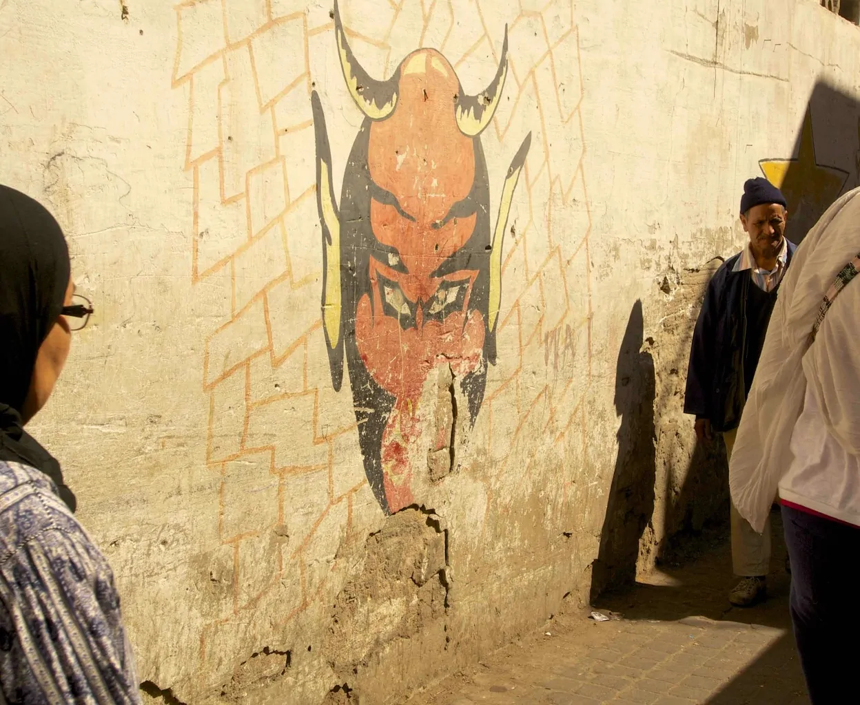 A woman in a hijab looking at a street art painting of the devil on a crumbling wall in Casablanca. 
