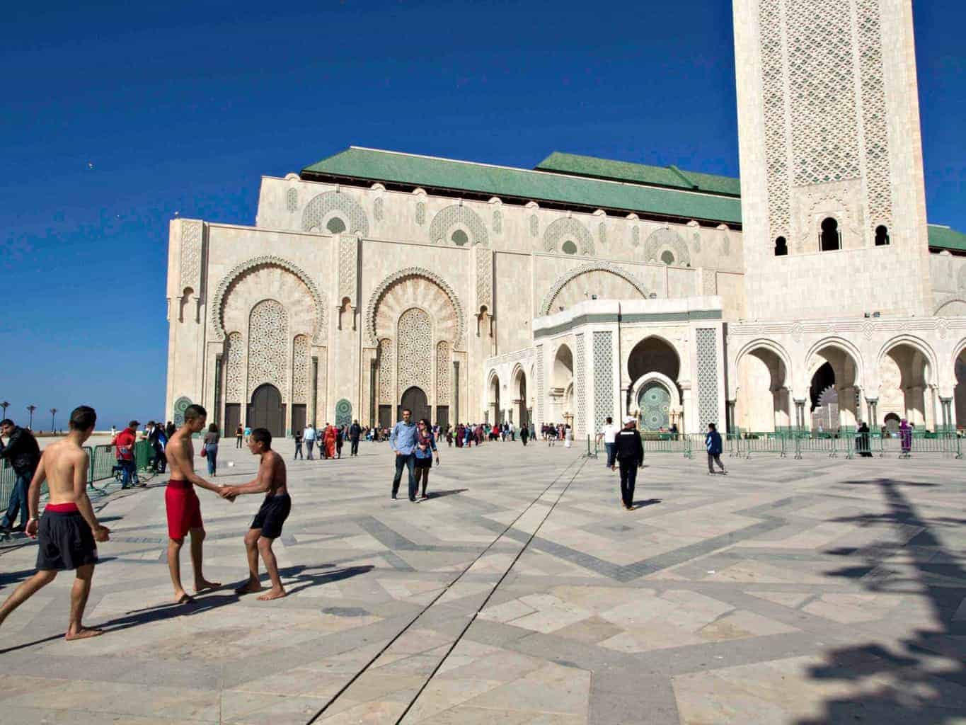 The outside of the Hassan II Mosque, with people wandering through the courtyard in front. 
